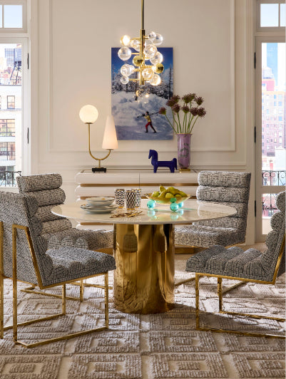 A luxury dining room featuring a round marble-top table with a chunky gold tree trunk base, surrounded by four upholstered dining chairs with gray and white textured patterns and gold frames. The room is decorated with a modern glass globe chandelier, a console table with a painting, and a patterned rug.