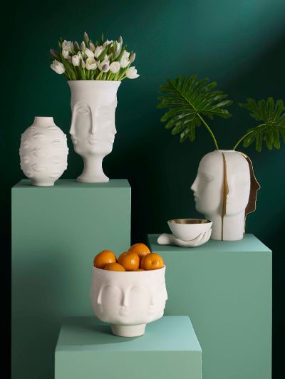 A styled interior scene with sculptural white ceramic head-shaped vases and bowls arranged on mint green pedestals against a dark teal wall. One vase holds white tulips, another contains orange fruits, and a third head-shaped vessel is shown in profile with a gold vertical accent and green leaves behind it.