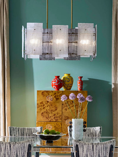 A modern four-light linear chandelier with frosted rectangular glass shades hangs over a dining table in a styled room with a teal wall, patterned chairs, and decorative vases.