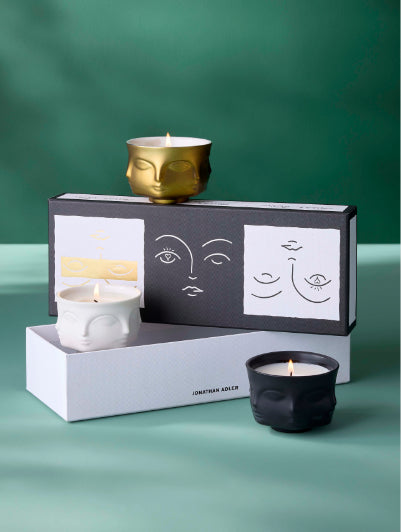 Jonathan Adler sculptural face-shaped candle holders in white, black, and gold, displayed with their branded packaging against a green background.