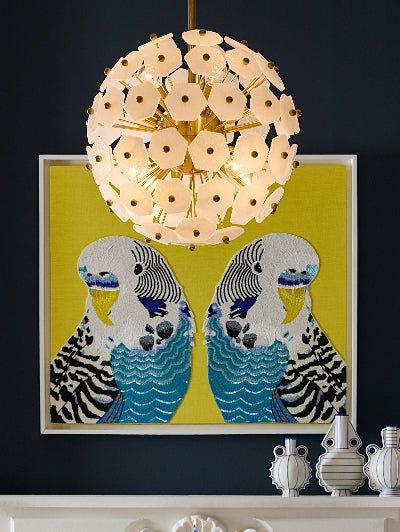 A decorative wall art piece featuring two stylized blue-and-white birds on a bright yellow background, hung on a dark navy wall beneath a circular pendant light. Small white ceramic vases sit on a white mantel below.