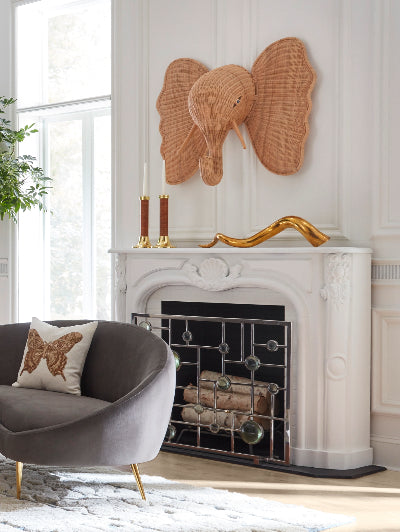 A large woven rattan elephant head wall decor piece mounted above a white fireplace mantel in a bright, modern living room. A gray velvet armchair with gold legs and a decorative pillow is visible in the foreground.