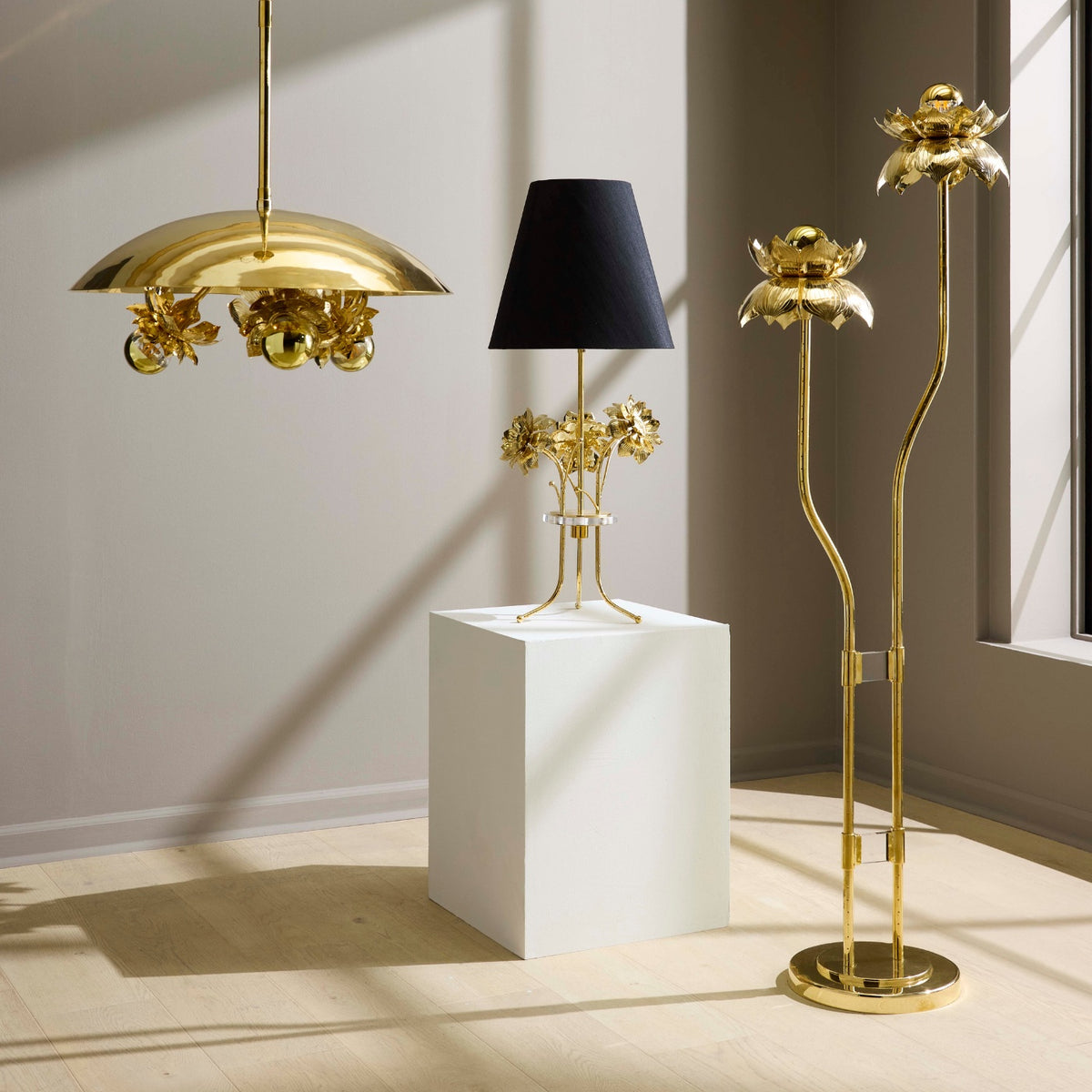 Gold floral-themed lamps and a black lampshade on a white cube in a room with sunlight casting shadows.