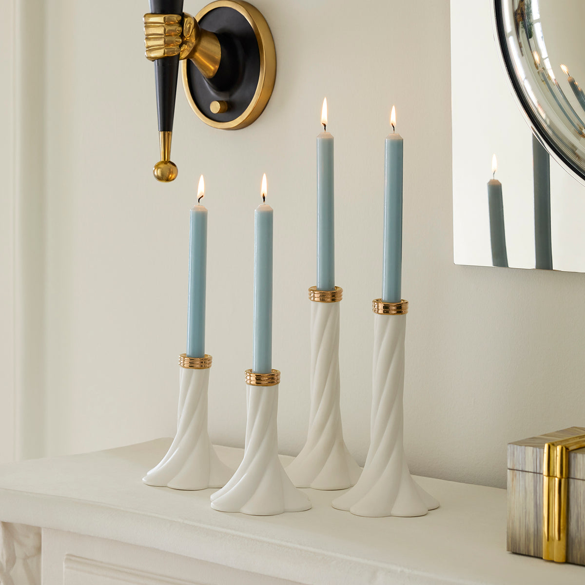 Four white twisted candlestick holders with gold accents holding light blue taper candles, styled on a white mantelpiece against a cream wall with a wall sconce and mirror visible.