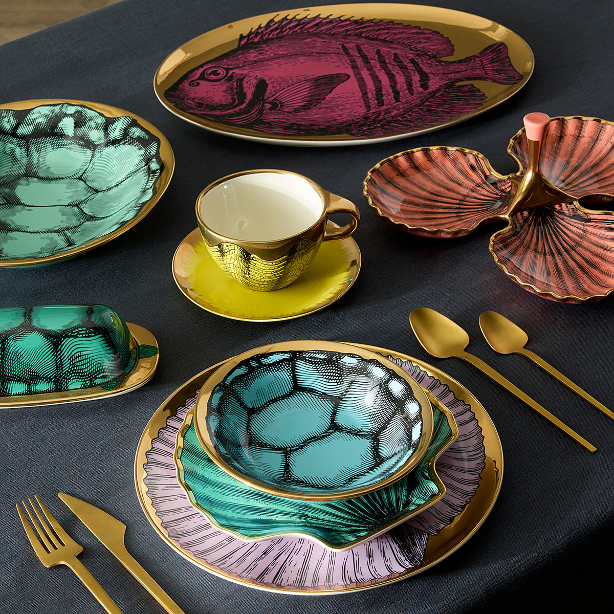 A styled tabletop dining set with gold-tone cutlery and ceramic tableware featuring shell and fish illustrations. The set includes plates, bowls, a cup and saucer, and serving dishes in teal, purple, pink, and gold on a dark tablecloth.
