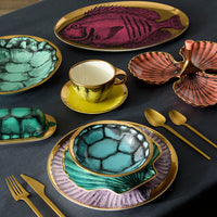 A styled tabletop dining set with gold-tone cutlery and ceramic tableware featuring shell and fish illustrations. The set includes plates, bowls, a cup and saucer, and serving dishes in teal, purple, pink, and gold on a dark tablecloth.