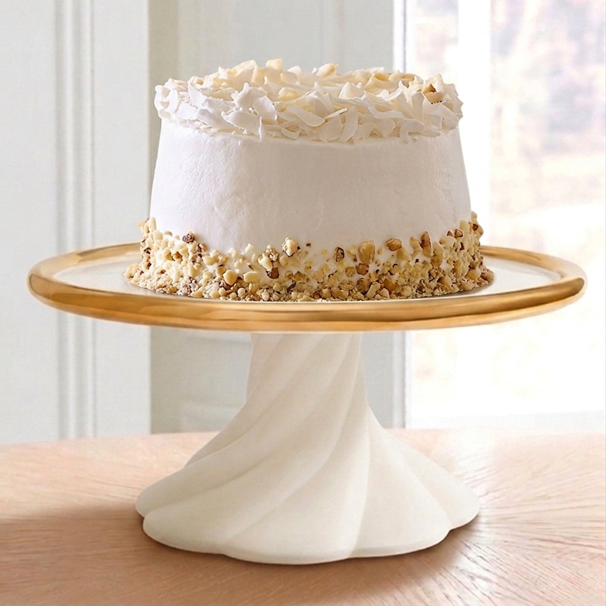 A white frosted cake with chopped nuts around the base sits on a white pedestal cake stand with a gold rim, placed on a wooden table in front of a softly blurred indoor background.