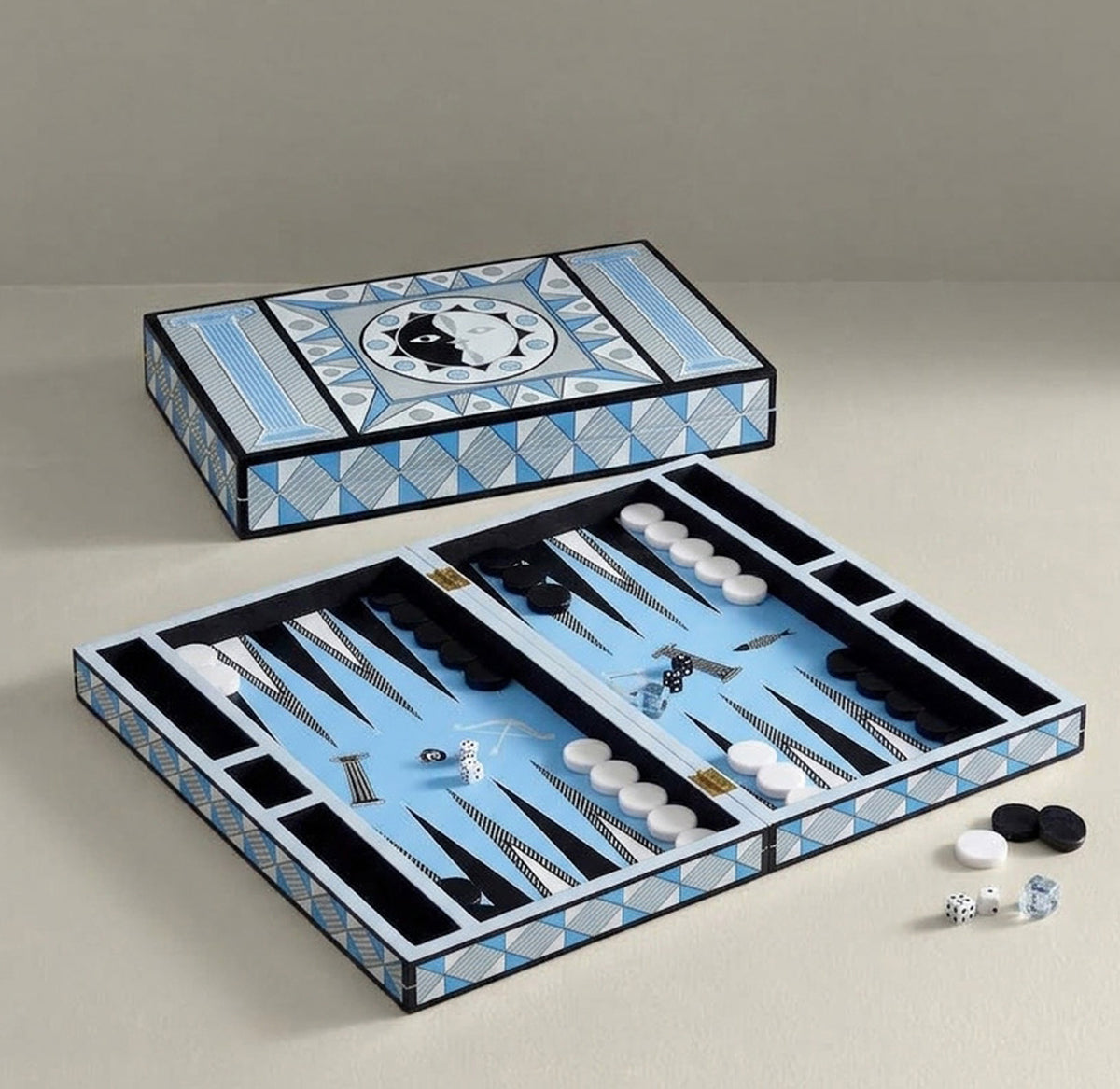 An open and closed backgammon set displayed on a neutral beige background. The game box features a geometric pattern in light blue, white, and black, and the open tray shows light blue felt playing surfaces with black and white checkers and pipettes.
