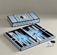 An open and closed backgammon set displayed on a neutral beige background. The game box features a geometric pattern in light blue, white, and black, and the open tray shows light blue felt playing surfaces with black and white checkers and pipettes.