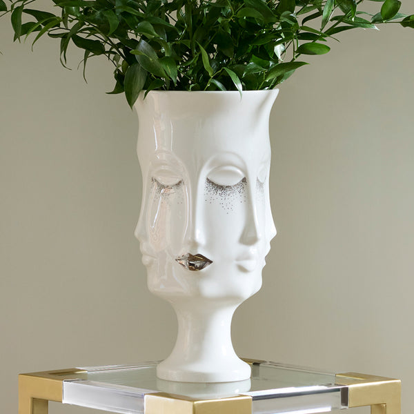 A white ceramic planter shaped like a stylized face with closed eyes and metallic silver glitter details, holding a green leafy plant, displayed on a gold-framed glass side table against a beige wall.