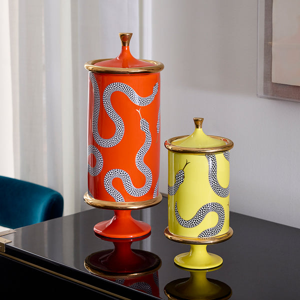 Two decorative lidded pedestal vases with snake motifs, one orange and one yellow, displayed on a glossy black table against a light gray wall.