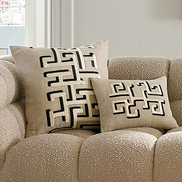 Two beige throw pillows with black geometric Greek key patterns resting on a textured beige sofa in a living room setting.