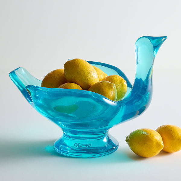A translucent turquoise blue glass bowl shaped like a duck, filled with yellow lemons, on a clean white background with two lemons resting beside it.