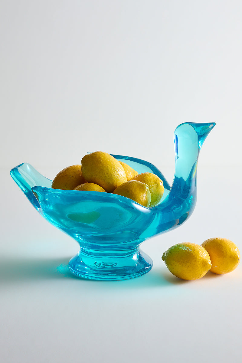 A translucent turquoise blue duck-shaped glass bowl filled with yellow lemons, with two additional lemons resting beside it on a clean white studio background.