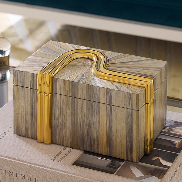 A rectangular decorative box with a striped wood-grain pattern in beige, brown, and gray, accented by a curved polished gold handle and gold side bands, resting on a book.