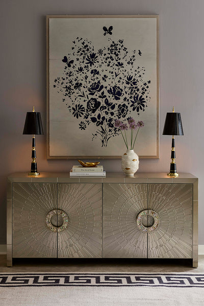 A styled interior scene featuring a large framed black floral artwork on a light gray wall above a metallic silver credenza, with two black table lamps, a small vase of flowers, and a patterned rug.