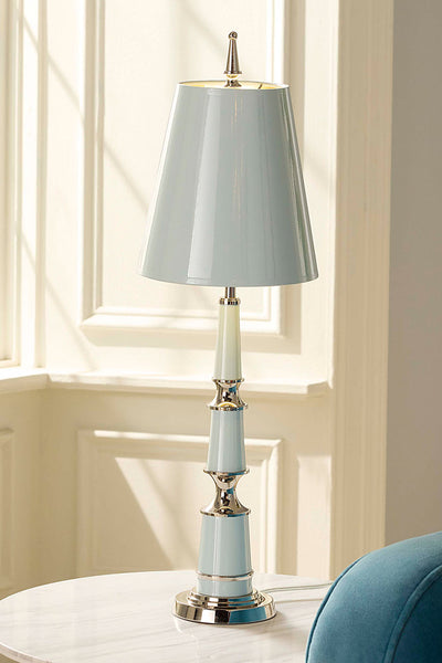 A table lamp with a light blue-gray shade and matching base, accented with polished gold-toned metal and a white ceramic neck, displayed on a white marble table in a sunlit room.