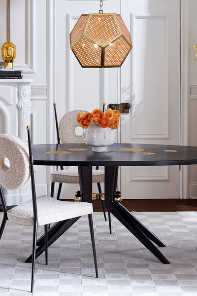A modern dining room scene featuring a round black table with gold leaf accents, textured cream boucle chairs, a geometric gold pendant light, and a white floral centerpiece on a patterned rug.