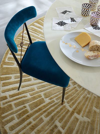 A modern teal velvet dining chair with a curved backrest and thin dark metal legs, positioned next to a round white table on a textured beige and cream brick-patterned rug.