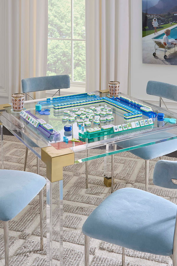 A modern glass-top dining table with a mahjong game set on top, surrounded by light blue upholstered chairs in a bright, contemporary dining room.
