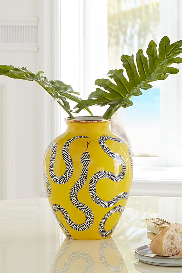 A bright yellow ceramic vase with a black and white snake pattern, holding green plant leaves, set on a reflective white table with a blurred kitchen background.