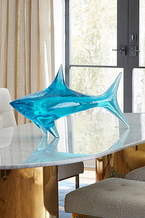 A translucent blue glass shark sculpture displayed on a marble-top dining table in a modern interior setting with neutral chairs and a window in the background.