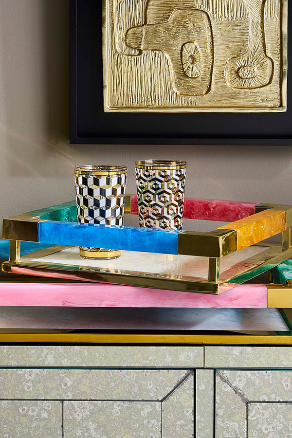A colorful decorative tray with gold metal edging and handles, styled on a mirrored cabinet with two patterned tumblers and framed wall art in the background.