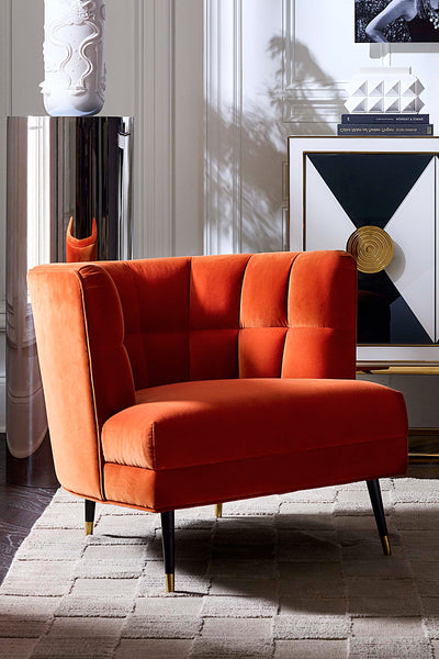 A modern orange velvet armchair with tufted back cushions and black tapered legs with gold tips, styled in a contemporary living room setting.