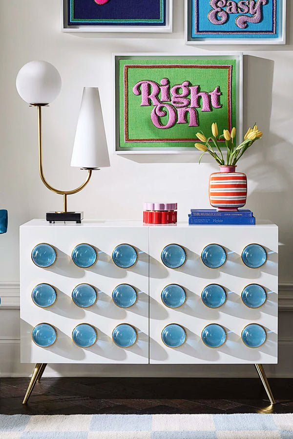 A white sideboard cabinet with gold legs and rows of round blue glass knobs, styled in a room setting with a lamp, books, a striped vase of tulips, and framed wall art.