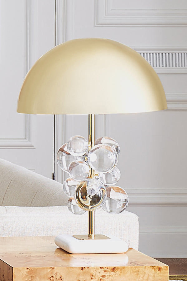 A modern table lamp with a matte gold dome shade, a gold-finished stem, clear glass bubble clusters, and a white square base, styled on a wooden side table in a bright room.