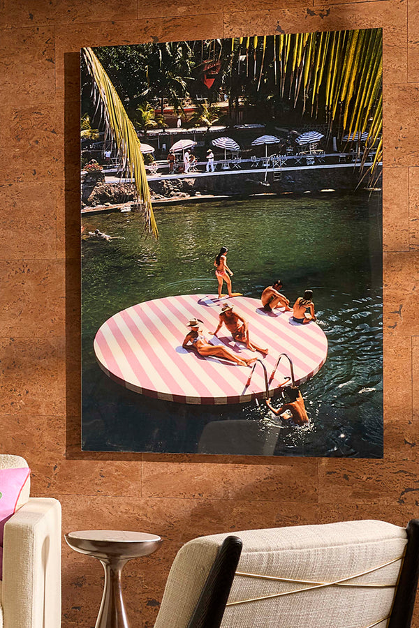 A large vertical wall art print depicting a resort pool scene with a pink-and-white striped floating platform, mounted on a textured brown wall above furniture.
