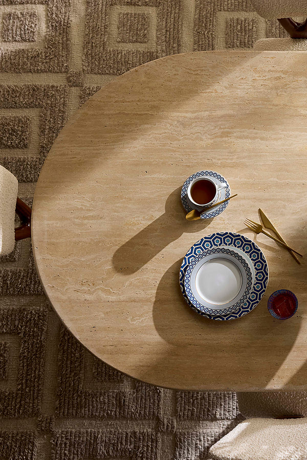 A top-down view of a round light wood table on a patterned rug, styled with a blue and white patterned teacup, a matching saucer, gold-toned cutlery, and a small red bowl.