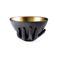 A decorative bowl with a matte black exterior and a metallic gold interior, shaped as if held from below by a sculpted hand, shown on a plain white background.