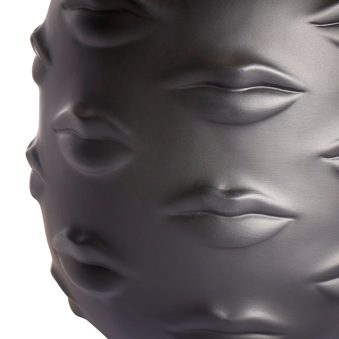 Close-up of a glossy dark gray sculptural object featuring embossed lips and eyes, photographed against a white background.