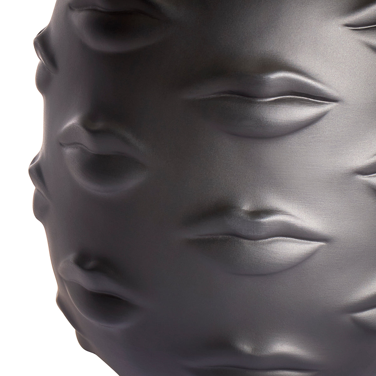 Close-up of a glossy dark gray sculptural object featuring embossed lips and eyes, photographed against a white background.