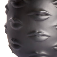 Close-up of a glossy dark gray sculptural object featuring embossed lips and eyes, photographed against a white background.
