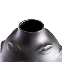 Close-up of a sculptural metallic vase or vessel with a wide circular opening, featuring embossed facial features including closed eyes and lips, in a dark silver finish against a white background.