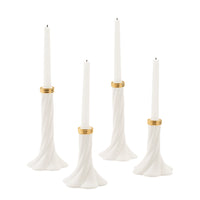 Four white tapered candles in sculptural flared holders with gold bands, arranged on a clean white background.