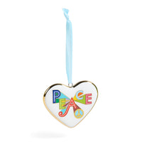 A heart-shaped white ornament with a gold-toned border, featuring the word 'PEACE' in colorful retro letters and a peace sign, hanging from a light blue ribbon on a white background.